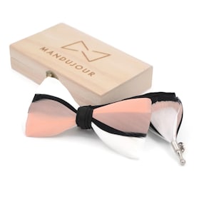 Puede incluir: Una pajarita de plumas rosa, negra y blanca con una cinta negra y un cierre plateado. La pajarita está en una caja de madera con la palabra "MANDUJOUR" grabada en la parte superior.