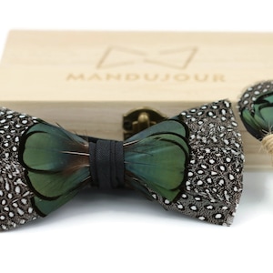 Puede incluir: Un pajarita de plumas verde y negra y un boutonnière a juego en una caja de madera con la palabra "MANDUJOUR" grabada en la tapa.