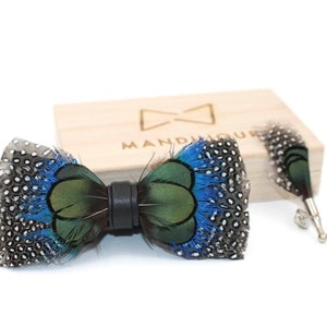 Puede incluir: Un pajarita de plumas azul y verde con una correa de cuero negro. La pajarita está en una caja de madera con la palabra "MANDULIQUE" grabada en ella.