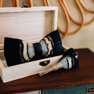 May include: A black and white feather bow tie with a leather strap in a wooden box. A matching feather pin is also included.