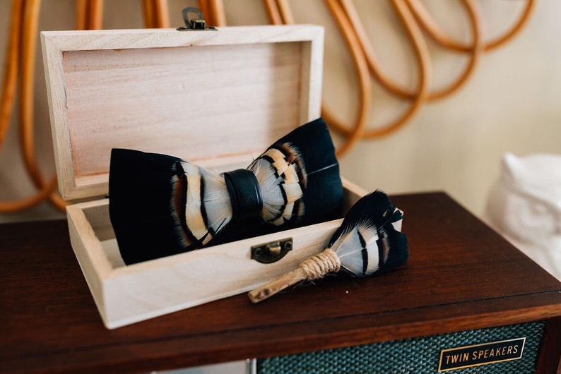 May include: A black and white feather bow tie with a leather strap in a wooden box. A matching feather pin is also included.