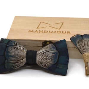 May include: A blue and brown feather bow tie with a black ribbon and a matching feather pin in a wooden box with the logo "MANDUJOUR" engraved on the lid.