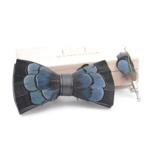 May include: A black bow tie with blue feathers and a matching feather lapel pin. The bow tie and pin are displayed in a wooden box.