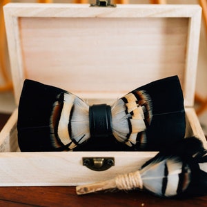 May include: A black bow tie with white and brown feathers, and a matching feather boutonniere, are displayed in a wooden box.