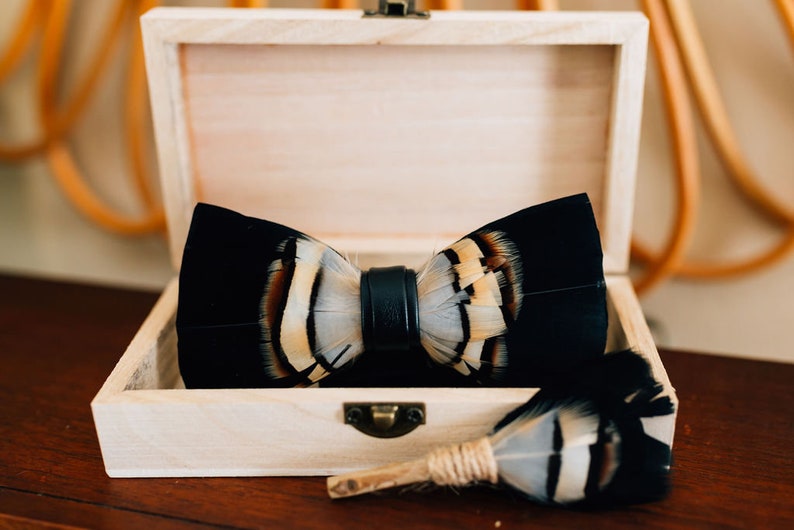 May include: A black bow tie with white and brown feathers, and a matching feather boutonniere, are displayed in a wooden box.