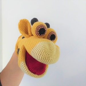 Op de afbeelding: Een gele en bruine gehaakte giraffenpop met een rode mond en zwarte ogen. De pop zit op de hand van een persoon.