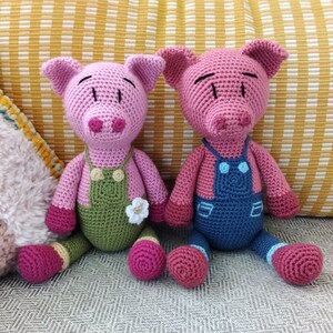 May include: Two crocheted pig plushies, one wearing a green overall and the other wearing a blue overall. Both plushies have pink bodies and black eyes.