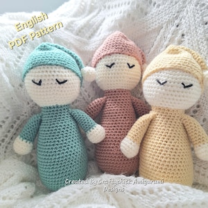 Puede incluir: Tres muñecas de ganchillo hechas a mano en colores pastel: turquesa, rosa y amarillo. Cada muñeca tiene una cara blanca con los ojos cerrados, un gorro a juego y un cuerpo sencillo. Las muñecas están sobre una superficie blanca texturizada. El texto "English PDF Pattern" es visible.