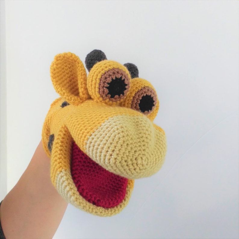 May include: A yellow and brown crocheted giraffe puppet with a red mouth and black eyes. The puppet is on a person's hand.