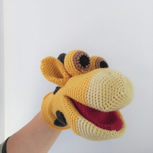 May include: A yellow and beige crocheted hand puppet shaped like a giraffe with black eyes and a red mouth.
