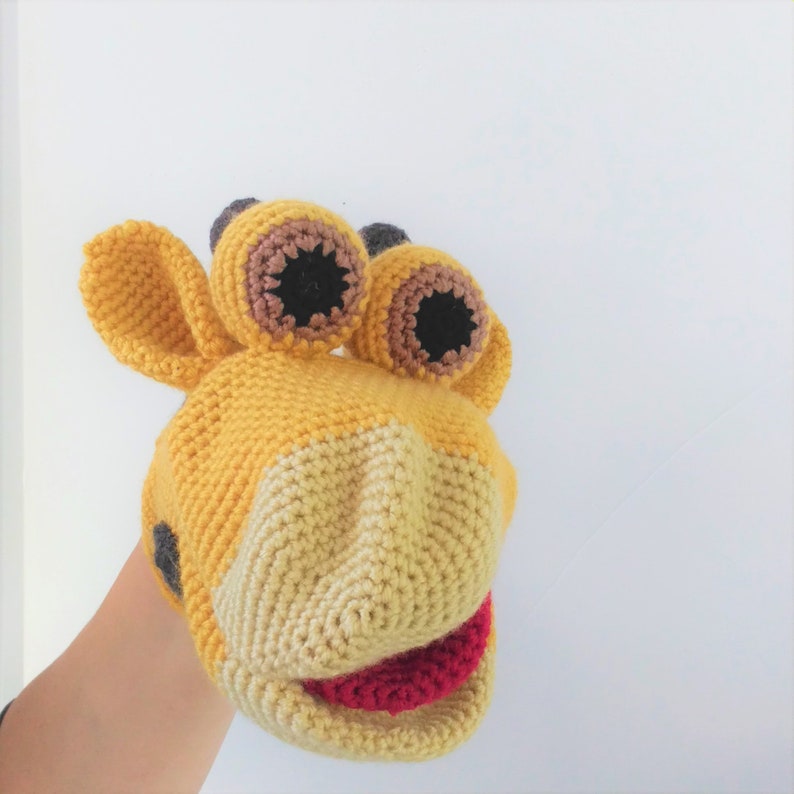 May include: A yellow crocheted puppet with brown and black eyes and a red mouth. The puppet is shaped like a giraffe and has a long neck and a small head.