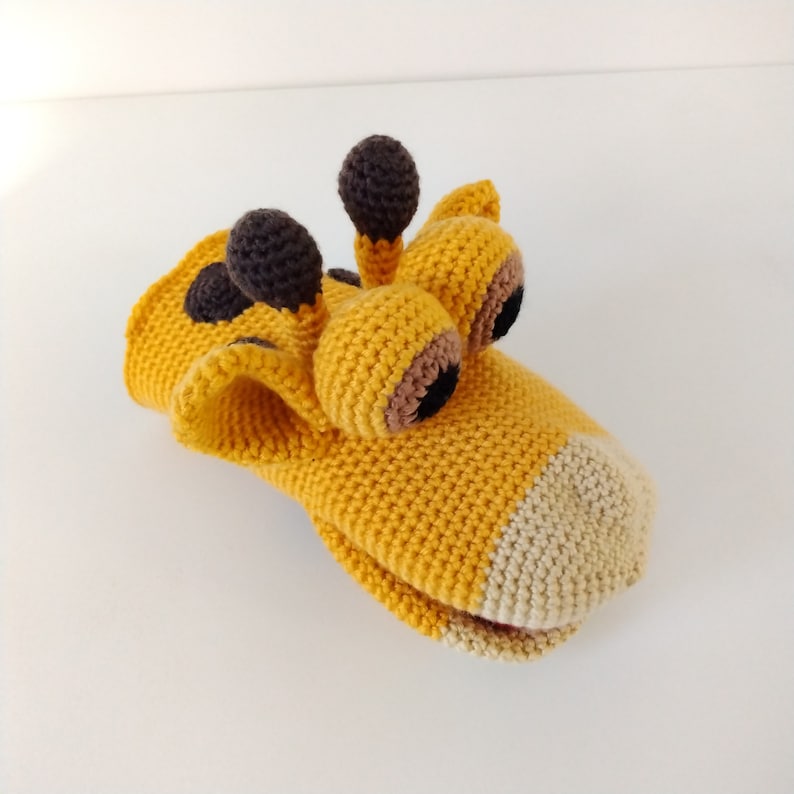May include: A crocheted giraffe puppet with brown eyes and a beige mouth. The puppet is made of yellow yarn and has a textured surface.