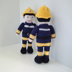 Pode incluir: Dois brinquedos de pelúcia de crochê vestidos de bombeiros. Ambos os brinquedos usam capacetes amarelos, calças listradas azul e amarelo e jaquetas azuis com cintos pretos. Os brinquedos estão em pé sobre uma superfície branca.