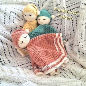 Puede incluir: Tres muñecos de apego para bebés tejidos a ganchillo en amarillo, turquesa y rosa, cada uno con una cara dormida y gorro a juego. El muñeco rosa está sobre una manta a juego con rayas blancas. Texto: "English PDF Pattern" y "Created By Craftychick Amigurumi Designs".