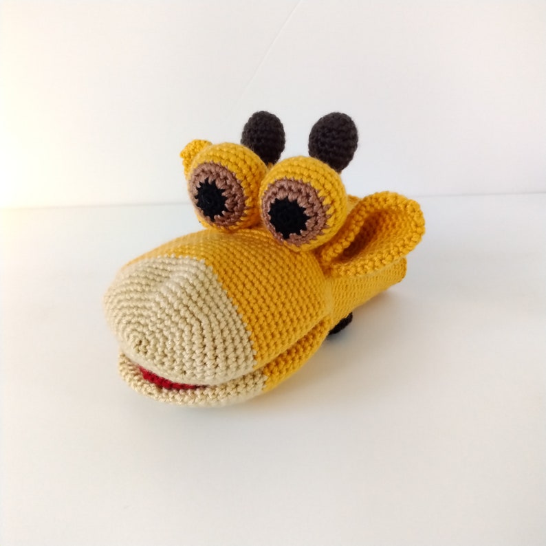 May include: A yellow and beige crocheted puppet with large brown eyes and a red mouth. The puppet is shaped like a giraffe and has a hand opening in the back.