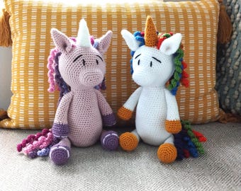 May include: Two handmade crochet unicorn dolls. One is light pink with a white horn and purple and pink mane and tail. The other is white with a rainbow mane and tail and an orange horn. Both have orange hooves.