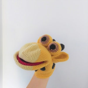 May include: A yellow and brown crocheted giraffe puppet with black eyes and a red mouth. The puppet is on a hand.