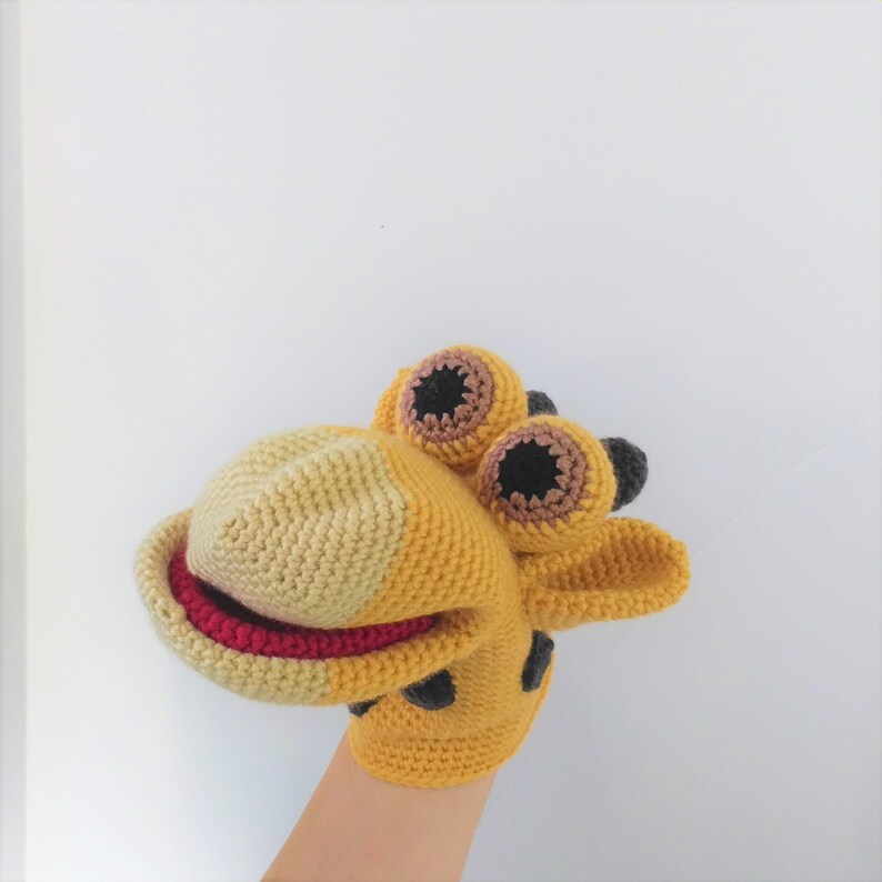 May include: A yellow and brown crocheted giraffe puppet with black eyes and a red mouth. The puppet is on a hand.