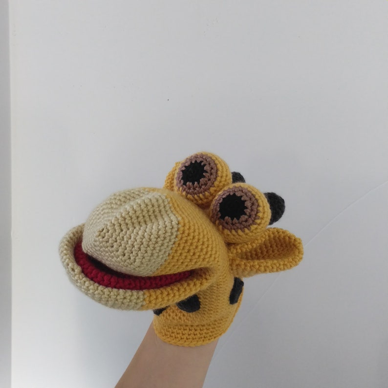 May include: A crocheted giraffe puppet with brown and black eyes, a red mouth, and a yellow and beige body.