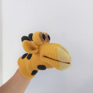 May include: A yellow crocheted giraffe puppet with black spots and brown eyes. The puppet has a large mouth that can be opened and closed.