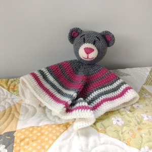May include: A gray crocheted teddy bear with pink ears and nose is attached to a white, pink, and gray striped crocheted baby blanket.