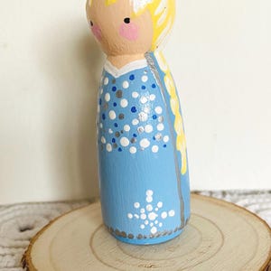 May include: A hand-painted wooden peg doll in a light blue dress with white and silver polka dots and a snowflake design. The doll has blonde hair and is displayed on a wooden slice.