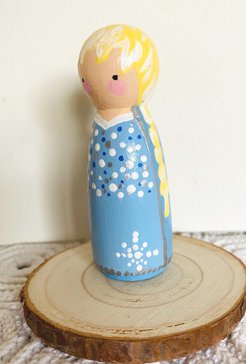 May include: A hand-painted wooden peg doll in a light blue dress with white and silver polka dots and a snowflake design. The doll has blonde hair and is displayed on a wooden slice.