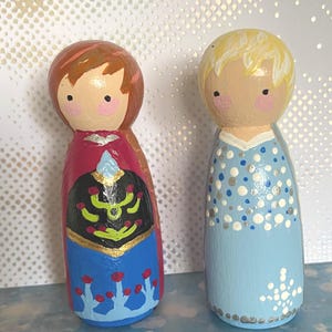 May include: Two hand-painted wooden peg dolls. One doll is dressed in a red, black, and blue outfit with gold accents. The other doll is in a light blue dress with silver and white details. Both dolls have painted faces with rosy cheeks.