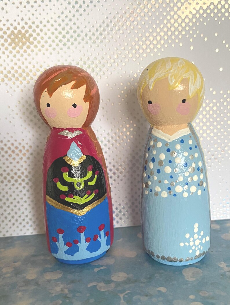 May include: Two hand-painted wooden peg dolls. One doll is dressed in a red, black, and blue outfit with gold accents. The other doll is in a light blue dress with silver and white details. Both dolls have painted faces with rosy cheeks.