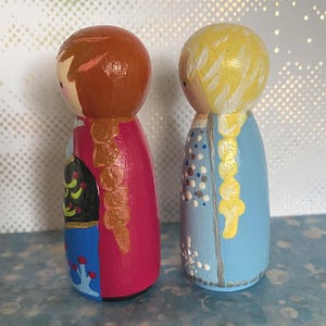 May include: Two hand-painted wooden peg dolls. One doll has brown hair and a pink dress, the other has blonde hair and a blue dress. The dolls feature painted details such as braids and floral patterns, giving a whimsical, handcrafted feel.