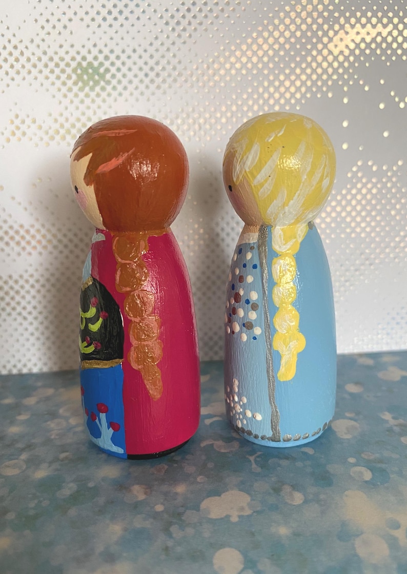 May include: Two hand-painted wooden peg dolls. One doll has brown hair and a pink dress, the other has blonde hair and a blue dress. The dolls feature painted details such as braids and floral patterns, giving a whimsical, handcrafted feel.