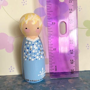 May include: A hand-painted wooden peg doll in a light blue dress with white and silver dot and floral details. The doll has blonde hair and rosy cheeks. A purple ruler is next to the doll, measuring approximately 7.6 cm.