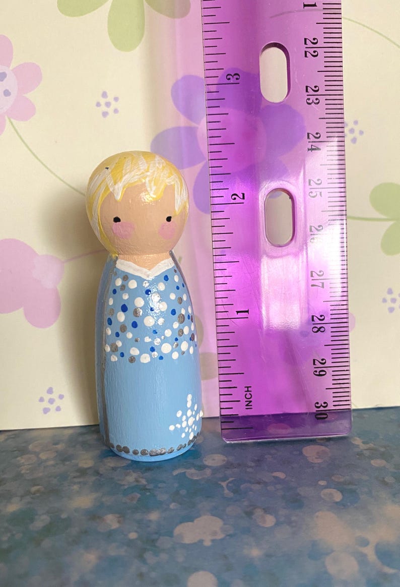 May include: A hand-painted wooden peg doll in a light blue dress with white and silver dot and floral details. The doll has blonde hair and rosy cheeks. A purple ruler is next to the doll, measuring approximately 7.6 cm.