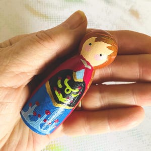 May include: Hand-painted wooden peg doll with a round head and a painted face. The doll is wearing a red top, blue skirt, and a black and gold design on the front. The doll is held in a hand.