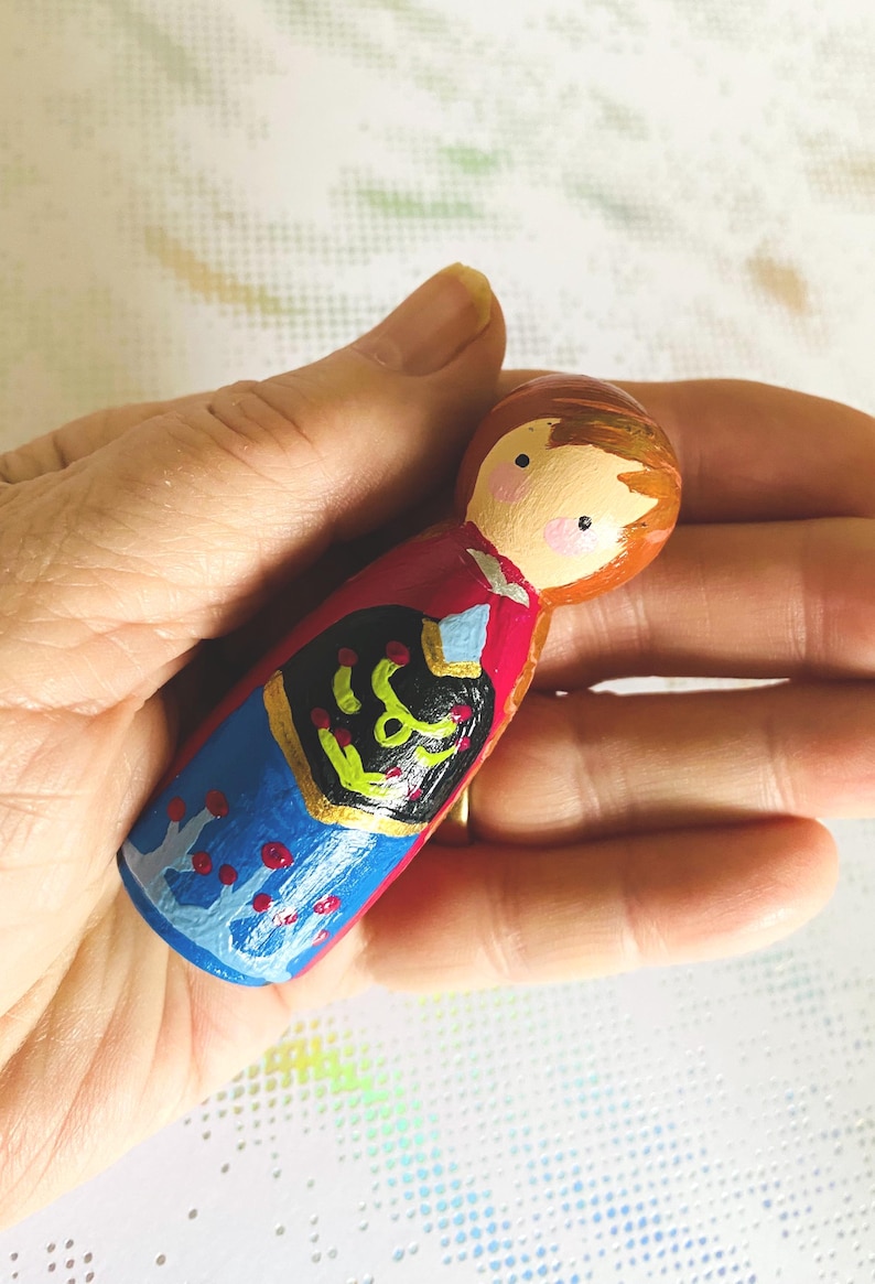 May include: Hand-painted wooden peg doll with a round head and a painted face. The doll is wearing a red top, blue skirt, and a black and gold design on the front. The doll is held in a hand.