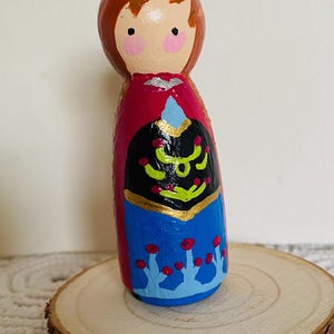 May include: A hand-painted wooden peg doll of a character with red hair, pink cheeks, and a red cape. The doll's dress is black with gold accents and blue with red floral details. The doll is displayed on a wooden slice.