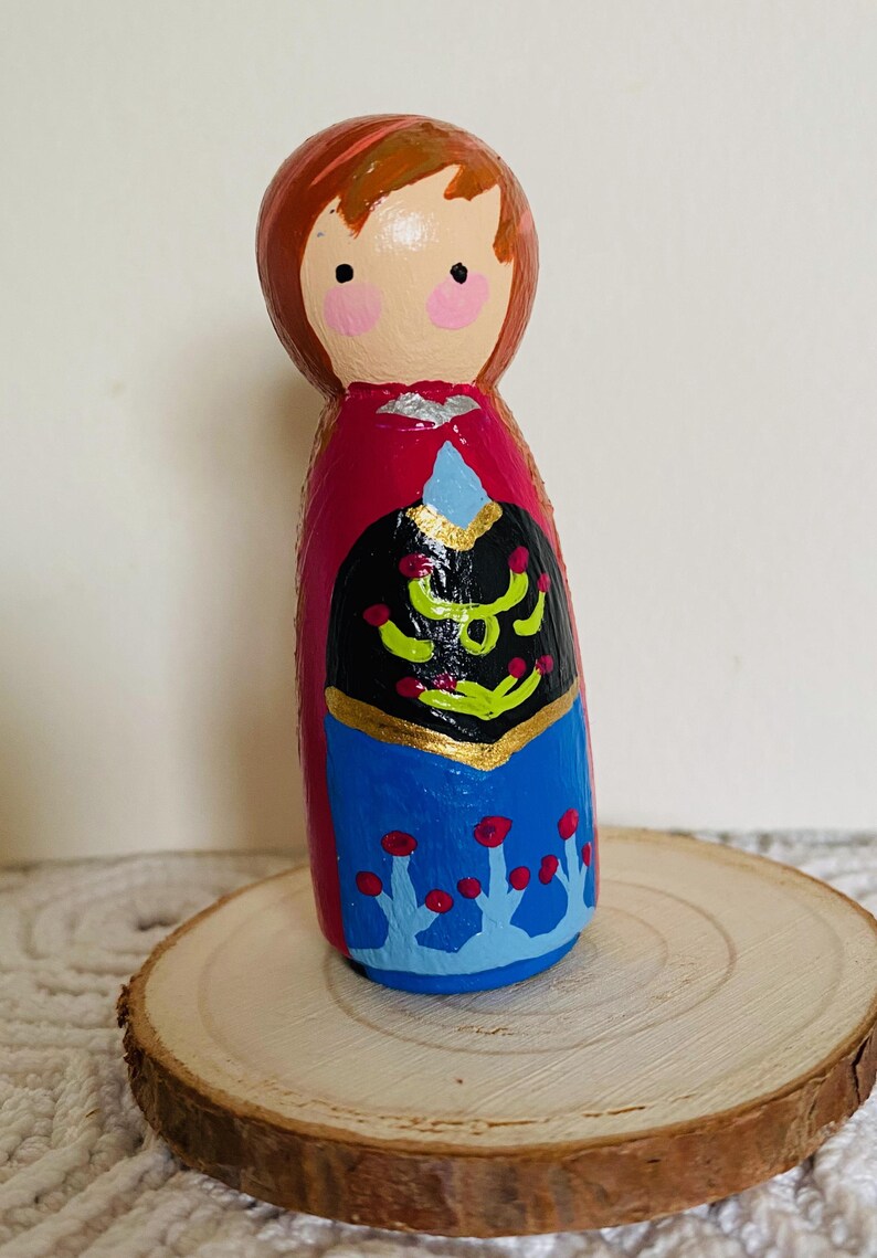 May include: A hand-painted wooden peg doll of a character with red hair, pink cheeks, and a red cape. The doll's dress is black with gold accents and blue with red floral details. The doll is displayed on a wooden slice.