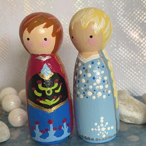 May include: Two hand-painted wooden peg dolls depicting cartoon characters. One doll has brown hair and a red and blue dress. The other has blonde hair and a light blue dress with white and silver details. The dolls are standing upright, side by side.