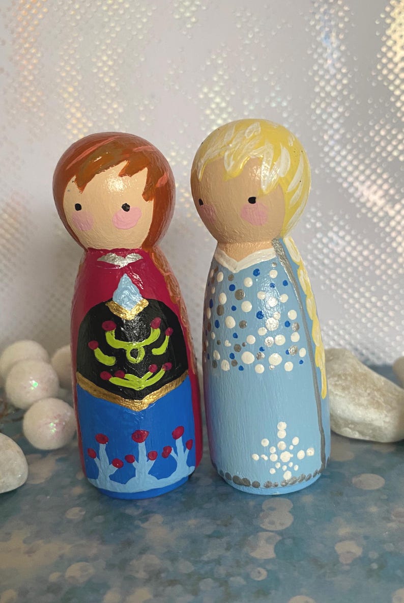 May include: Two hand-painted wooden peg dolls depicting cartoon characters. One doll has brown hair and a red and blue dress. The other has blonde hair and a light blue dress with white and silver details. The dolls are standing upright, side by side.