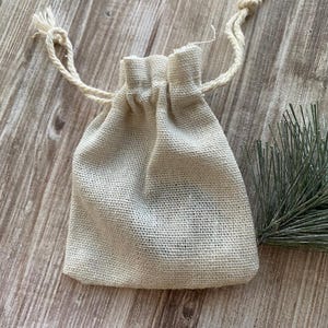 May include: A small, beige burlap drawstring bag with a woven texture. The bag is gathered at the top with a natural-coloured cord, and the ends of the cord are knotted with tassels. A sprig of green foliage is in the background.