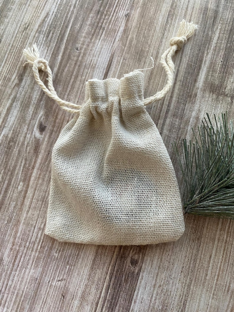 May include: A small, beige burlap drawstring bag with a woven texture. The bag is gathered at the top with a natural-coloured cord, and the ends of the cord are knotted with tassels. A sprig of green foliage is in the background.