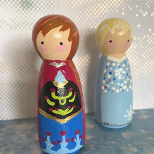 May include: Two hand-painted wooden peg dolls. One doll is dressed in a red, black, and blue outfit with gold accents. The other doll is wearing a light blue dress with white polka dots. Both dolls have painted faces with rosy cheeks.