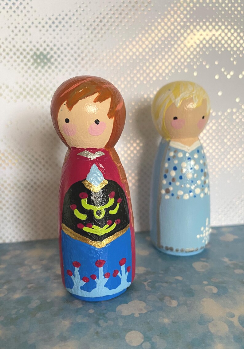 May include: Two hand-painted wooden peg dolls. One doll is dressed in a red, black, and blue outfit with gold accents. The other doll is wearing a light blue dress with white polka dots. Both dolls have painted faces with rosy cheeks.