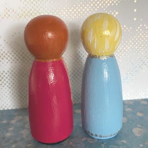 May include: Two wooden peg dolls. One doll has a copper-coloured head and a magenta body. The other doll has a yellow head and a light blue body. Both dolls have a gold-coloured band at the neck and are on a blue surface.