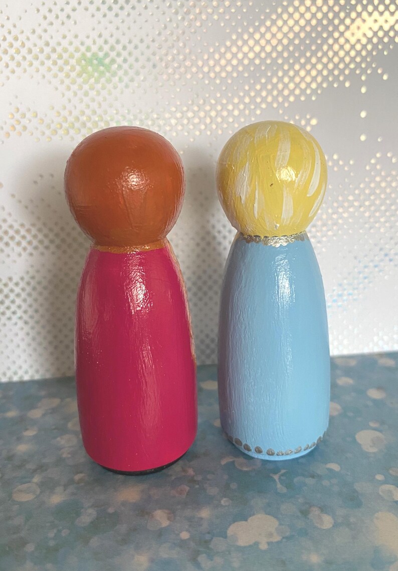 May include: Two wooden peg dolls. One doll has a copper-coloured head and a magenta body. The other doll has a yellow head and a light blue body. Both dolls have a gold-coloured band at the neck and are on a blue surface.