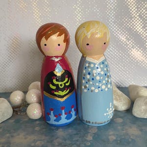 May include: Two hand-painted wooden peg dolls. One doll has red hair and a red, black, and blue dress. The other doll has blonde hair and a light blue dress with white and silver polka dots. The dolls are standing next to each other.