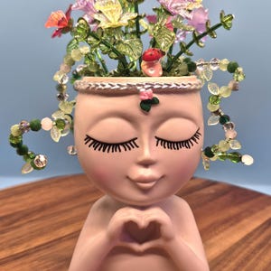 May include: A light pink planter shaped like a head with closed eyes and long eyelashes, featuring hands forming a heart shape. The planter is filled with colorful glass flowers, green stems, and a small red mushroom, with beaded vines draped over the sides.