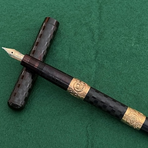 Puede incluir: Una pluma estilogr&aacute;fica vintage con plum&iacute;n de oro y bandas doradas ornamentadas. El cuerpo de la pluma es negro con un patr&oacute;n de cuadros, y la tapa est&aacute; separada. La pluma descansa sobre una superficie verde.