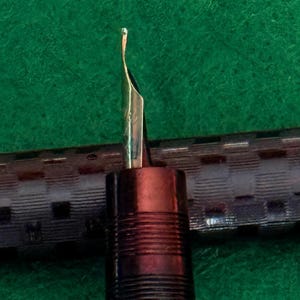 Puede incluir: Primer plano de una pluma estilogr&aacute;fica con punta dorada. El cuerpo de la pluma es marr&oacute;n oscuro con ranuras horizontales. El fondo es una superficie verde texturizada.