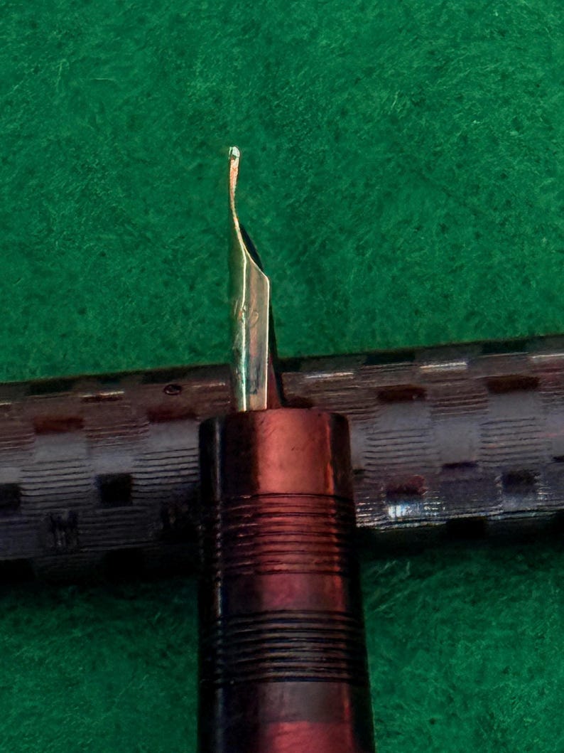 Puede incluir: Primer plano de una pluma estilogr&aacute;fica con punta dorada. El cuerpo de la pluma es marr&oacute;n oscuro con ranuras horizontales. El fondo es una superficie verde texturizada.